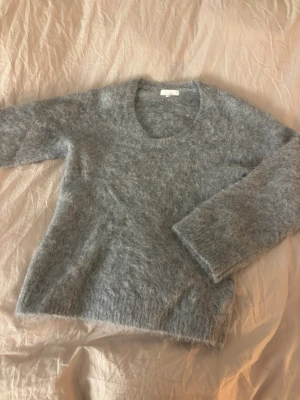 Ljusgrå fluffig stickad tröja - Supermjuk och fluffig ljusgrå alpackan tröja med rund halsringning och långa ärmar. Tröjan har en avslappnad passform och är gjord i ett lurvigt material som känns riktigt cozy. Perfekt för kalla dagar när du vill vara både varm och snygg.