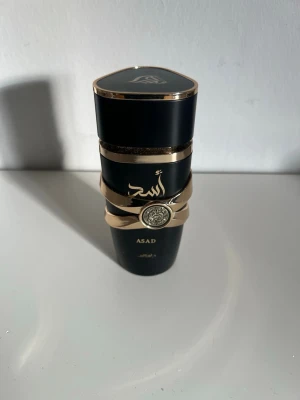 Asad parfym från Lattafa svart/guld  - Snygg parfymflaska från Lattafa i svart med gulddetaljer och arabiska tecken. Flaskan har en unik form med dekorativt mönster och metallring runt mitten. Perfekt för dig som gillar exklusiva och stilrena dofter. Lite mer än halva kvar!