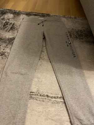 Grå mjukisbyxor Tommy Hilfiger - Snygga grå mjukisbyxor från Tommy Hilfiger med logga och text längs ena benet. Byxorna har dragsko i midjan och är tillverkade i mjukt bomullsmaterial. Perfekta för en avslappnad och trendig stil.