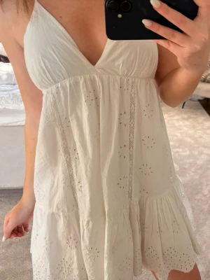Vit somrig klänning med spetsdetaljer - Söt vit kortklänning med v-ringning och tunna axelband. Klänningen har broderade spetsdetaljer och ett luftigt, volangigt fall. Perfekt för varma dagar och har en öppen rygg för extra sommarkänsla.
