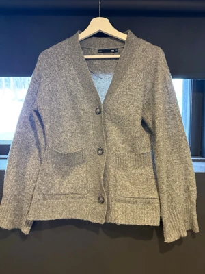 Grå kofta med fickor - Mysig grå kofta från med två stora fickor framtill och knäppning med tre knappar. Koftan har ribbade muddar och en avslappnad passform, perfekt att slänga på sig när det är kyligt. Tillverkad i mjukt stickat material.