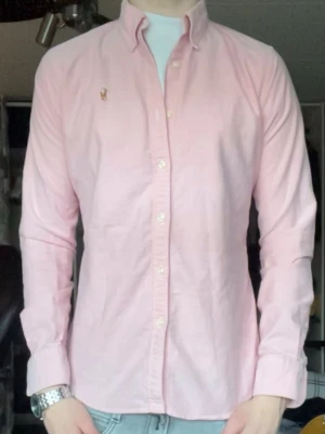 Skjorta - Klassisk rosa skjorta från Ralph Lauren med button-down krage och broderad logga på bröstet. Skjortan har långa ärmar, knäppning framtill och är tillverkad i bomull. Perfekt för en stilren och avslappnad look.