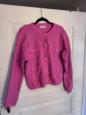 Rosa kofta från Holly & Whyte - Säljer en rosa kofta från Holly & Whyte med rund halsringning och dekorativa guldknappar framtill. Koftan har två bröstfickor och två fickor nertill, alla med knappar. Grovstickad struktur och lång ärm, perfekt för att liva upp din outfit.