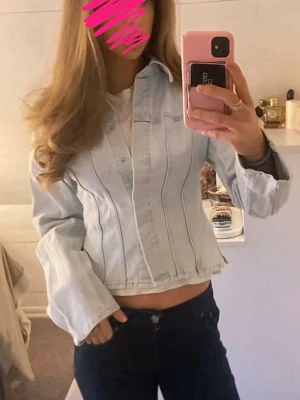 Jeans tröja  - Ljusblå croppad jeansjacka med klassisk krage och långa ärmar. Jackan har markerade sömmar framtill och knäppning längs hela framsidan. Perfekt för en trendig och avslappnad stil.