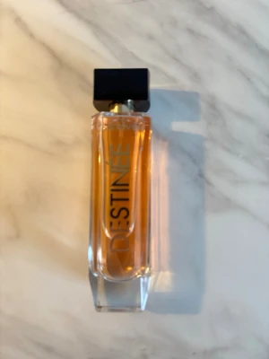 La Rive Destinée Eau de Parfum - La Rive Destinée är en parfym i en elegant, rektangulär glasflaska med svart fyrkantigt lock och guldiga detaljer. Flaskan har en varm, bärnstensfärgad vätska och modern design med tydlig logotyp på framsidan. Perfekt för dig som gillar stilrena och trendiga dofter.