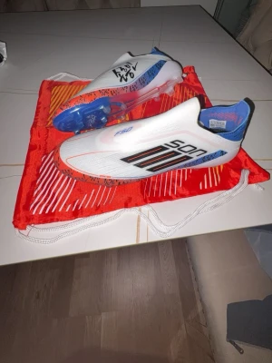 Adidas F50 Son fotbollsskor - Säljer ett par Adidas F50 fotbollsskor i vitt med blå och röda detaljer samt svarta SON-tryck på sidan. Skorna har en strömlinjeformad design utan snörning och genomskinlig dobbsula. Perfekta för dig som vill sticka ut på planen.