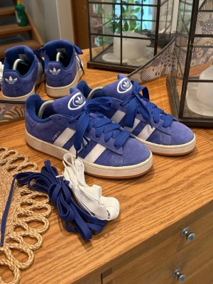 Blå Adidas Campus sneakers i mocka - Snygga blå Adidas Campus sneakers i mocka med vita detaljer och klassiska tre ränder på sidan. Skorna har vit sula, blå och vita skosnören samt Adidas logga på plös och häl. Perfekta för dig som gillar retrostil och streetwear.