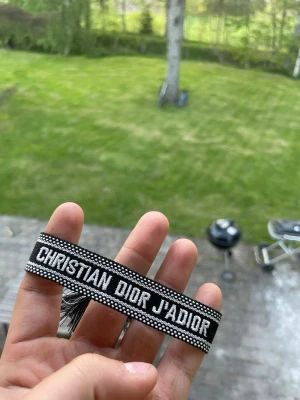 Christian Dior J'Adior armband svart - Snyggt svart tygarmband från Christian Dior med texten 'J'ADIOR' i vitt. Armbandet har en vävd design med vita detaljer längs kanterna och är justerbart för att passa de flesta handleder. Perfekt accessoar för att lyfta din outfit.