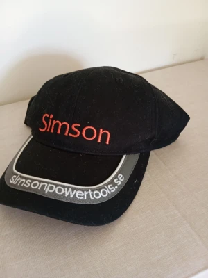 Svart keps från Simson - Svart keps med röd Simson-logga broderad framtill och texten simsonpowertools.se på skärmen. Klassisk form med böjd skärm och justerbar baksida. Perfekt accessoar för en sportig och avslappnad stil.