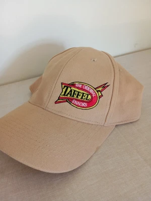 Beige keps Yafell Snacks brodyr - Snygg beige keps med broderad Yafell Snacks-logga i rött, gult och svart framtill. Klassisk böjd skärm och justerbar baksida. Perfekt accessoar för en avslappnad streetstyle-look.