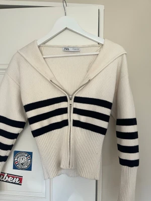 Vit stickad tröja med marinblå ränder Zara - Säljer en vit stickad tröja från Zara med marinblå horisontella ränder på både kropp och ärmar. Tröjan har dragkedja framtill, bred krage i sjömansstil och ribbstickade muddar. Perfekt för en trendig och avslappnad look.