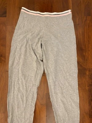 Grå loungebyxor från Calvin Klein - Mysiga grå loungebyxor från Calvin Klein med elastisk midja och rosa/vit logotypdetalj i midjebandet. Perfekta för chill hemma eller när du vill vara bekväm. Tillverkade i mjukt och stretchigt material.