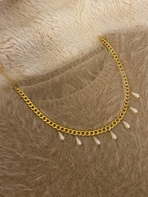 Guldfärgat halsband med pärldroppar - Snyggt halsband i guldfärgad rostfritt med kedja och sju hängande droppformade pärlor. Perfekt för att ge en extra touch till din outfit. Kedjan har en klassisk design och pärlorna ger en elegant känsla.