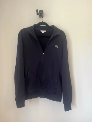 Mörkblå zip-hoodie från Lacoste - Mörkblå hoodie med dragkedja från Lacoste. Klassisk design med ståkrage, två fickor framtill och den ikoniska gröna krokodilloggan på bröstet. Perfekt för en avslappnad och sportig stil. Storlek S, skick 7/10 