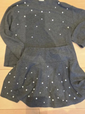 Grå stickad set med pärldetaljer - Grått stickat set med lång ärm och hög hals, dekorerat med små vita pärlor på både överdel och nederdel. Mjukt material och en lätt utställd kjol ger en mysig och trendig look.