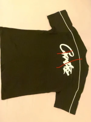 Corteiz Oversized - Cool svart t-shirt med vit grafisk text på bröstet och ryggen samt tunna vita linjer över axlarna. Klassisk passform och korta ärmar, perfekt till jeans eller shorts. Materialet känns mjukt och bekvämt mot huden.