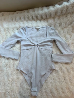 Ljusgrå ribbad body med knutdetalj - Säljer en ljusgrå ribbad body från H&M med lång ärm och snygg knutdetalj framtill under bysten. Bodyn har djup ringning och är tillverkad i ett mjukt, stretchigt material som sitter skönt mot kroppen.