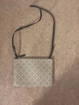 Beige axelväska med rutmönster - Snygg beige by malene birger  axelväska med vitt rutmönster och justerbar svart axelrem. Väskan har en rektangulär form och stängs med dragkedja. Insidan är fodrad i beige tyg. Perfekt för att bära det viktigaste med stil.