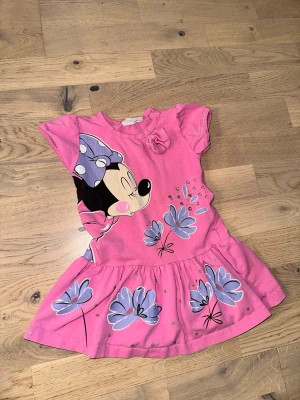 Rosa klänning med Musse Pigg-tryck - Rosa klänning med Musse Pigg-tryck Söt rosa klänning med kort ärm och stort tryck av Musse Pigg samt lila blommor. Kjolen är utställd och klänningen har en liten rosett vid halsen. Perfekt för dig som gillar färgglada och lekfulla plagg.