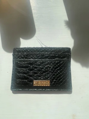 Svart korthållare By Bianca Ingrosso - Snygg svart korthållare i präglat läder från By Bianca Ingrosso. Plånboken har flera kortfack och en guldfärgad metallbricka med logga på framsidan. Perfekt för dig som vill ha ett stilrent och smidigt sätt att bära dina kort.