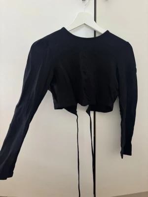 Svart croppad topp med snörning - Trendig svart croppad topp med långa ärmar och snörning som kan knytas runt midjan. Toppen har en enkel, stilren design och är perfekt för att skapa en edgy look. Materialet känns mjukt och lätt mot huden.