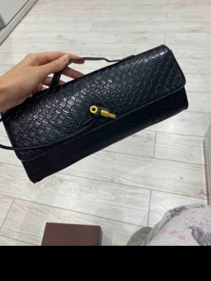 Svart kuvertväska med ormmönster - Snygg svart kuvertväska i konstläder med ormmönstrad struktur. Väskan har ett unikt guldfärgat låsdetalj framtill och ett smalt handtag upptill. Perfekt accessoar för att lyfta din outfit med en trendig touch.