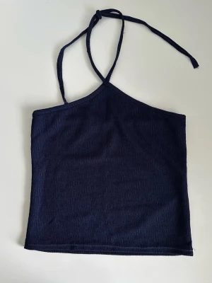 Mörkblått strukturerat halterneck linne - storlek S - Superfint halternecklinne / halterneck-topp i marinblått elastiskt tyg med diskret strukturerad textur. Smala justerbara knytband i nacken. Toppen är designad med en figurnära passform och en rak nederkant. Storlek S. Superfint skick, som ny! 
