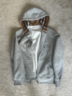 Grå zip-hoodie från Burberry - Snygg grå hoodie med dragkedja från Burberry. Klassisk huva med det ikoniska rutmönstret på insidan. Mjukt material och två fickor framtill. Perfekt för en stilren och avslappnad look. Helt oanvänd 