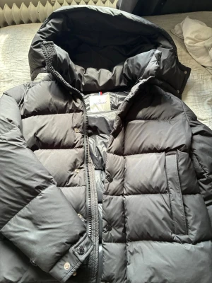 Svart dunjacka från Moncler - Säljer en svart dunjacka från Moncler med stor huva och quiltad design. Jackan har dragkedja och tryckknappar, samt praktiska sidofickor. Insidan har klassisk Moncler-etikett och serietidningsdetalj. Perfekt för kalla vinterdagar.