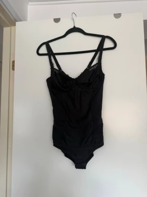 Svart body med spetsdetaljer - Snygg svart body med tunna axelband och vacker spets upptill vid bysten. Bodyn har bygel och är tillverkad i ett glansigt, stretchigt material som sitter tajt mot kroppen. Perfekt för att skapa en stilren och trendig look.