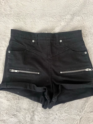 Svarta high waist shorts med dragkedjor - Snygga svarta high waist shorts med uppvikta ben och coola dragkedjor framtill. Shortsen har bälteshällor, fickor och en stor dragkedja bak. Perfekta för dig som gillar en edgy stil.