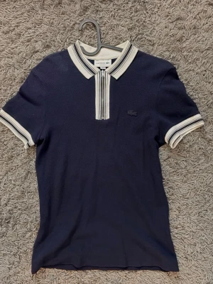 Marinblå polotopp från Lacoste - Snygg marinblå polotopp från Lacoste med vit krage och ärmslut, samt diskreta ränder. Dragkedja framtill och klassisk Lacoste-logga på bröstet. Perfekt för dig som gillar stilrena och sportiga plagg.
