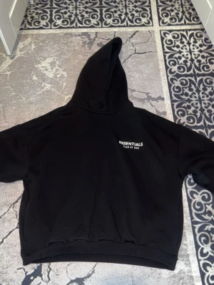 Svart Essentials Fear of God hoodie - Säljer en svart hoodie från Essentials Fear of God med vit logga på bröstet och stor text på ryggen. Hoodien har en klassisk huva och ribbade muddar vid ärmar och nederkant. Perfekt för dig som gillar streetwear och vill ha en stilren look. Pris går att diskutera. Annan färg finns samt byxor till den.