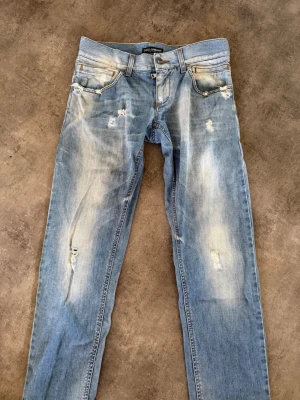 Ljusblå jeans Dolce & Gabbana - Säljer ett par ljusblå jeans från Dolce & Gabbana med slitningar och distressed detaljer. Jeansen har fem fickor, bälteshällor och klassisk knappgylf. Perfekt för en avslappnad och trendig look.
