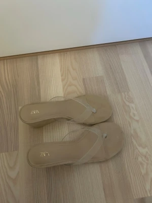 Beige kilklackade sandaler från Zara - Säljer ett par snygga beige sandaler från Zara med kilklack och transparenta remmar. Skorna har öppen tå och en modern, minimalistisk design som passar perfekt till sommarens outfits. Klacken ger extra höjd och remmarna är i plastmaterial.
