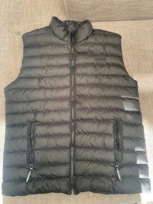 Svart dunväst från Hugo Boss - Snygg svart dunväst från Hugo Boss med diskret logga broderad på bröstet. Västen har ståkrage, två dragkedjeförsedda fickor och quiltad design. Perfekt att slänga över en hoodie eller tröja för extra värme. Materialet är glansigt och känns riktigt modernt.