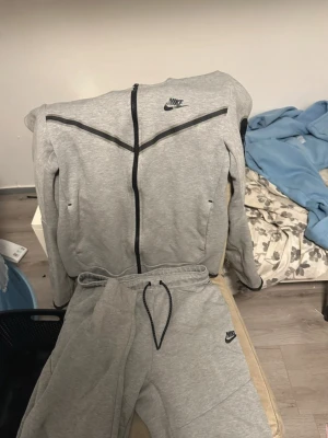 Grått Nike set - Säljer ett grått set från Nike med matchande zip-hoodie och byxor. Tröjan har svarta detaljer, dragkedja framtill och Nike-logga på bröstet. Byxorna har resår i midjan med snörning och Nike-logga på benet. Perfekt för en sportig och avslappnad stil. Koftan är i S och byxorna i M