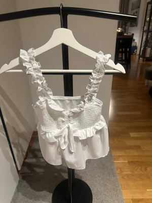 Vit volangtop med knyt och smock S - Supersöt vit topp med volangdetaljer och smockad överdel. Axelbanden är breda och rynkade för en extra gullig look. Toppen har snörning framtill och är perfekt för varma dagar.
