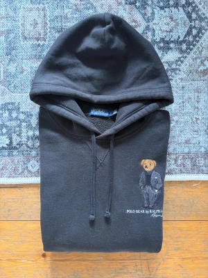 Svart Polo Ralph Lauren Bear Hoodie - Storlek M - Hej! 👋 Säljer en riktigt snygg och ikonisk svart Polo Ralph Lauren "Polo Bear"-hoodie i storlek M. 🐻✨ Ett måste för varje samling! Tröjan är i mycket gott skick och välvårdad. Det kan förekomma något enstaka litet nopprigt parti (helt naturligt för materialet), men det är inget som stör helhetsintrycket. 100% äkta med tvättlappar kvar (ingen QR-kod på denna modell). En riktig stilikon som passar till allt. Fraktar snabbt! 🚛📦💬💯 