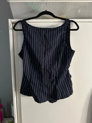 Mörkblå randig linnetopp med knyt - Snygg mörkblå linnetopp med vita vertikala ränder. Toppen har ärmlös design och ett coolt knyt i ryggen som ger en öppen rygg-look. Perfekt för dig som gillar stilrena detaljer och vill ha något luftigt till sommaren. Liten Office vibe
