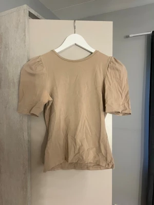 Beige topp med puffärm/sweter - Trendig beige topp med puffärmar och rund halsringning. Tillverkad i mjukt bomullsmaterial och har en enkel, stilren design som passar till många olika outfits. Perfekt för dig som gillar en clean och modern look.