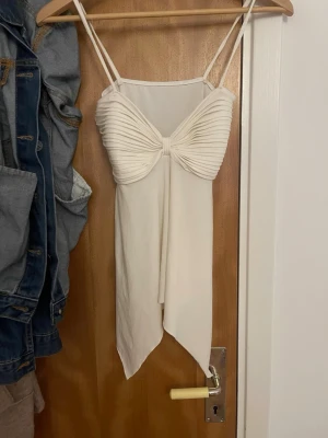 Vit linnetopp med draperad front - Snygg vit linnetopp med tunna axelband och draperad detalj framtill. Toppen har en asymmetrisk nederkant och en elegant twistad effekt över bysten. Perfekt för att skapa en trendig och stilren look.