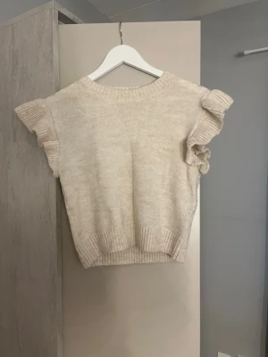 Beige stickad topp från Gina Tricot/sweter - Supersöt beige stickad topp från Gina Tricot med volangdetaljer på ärmarna och ribbstickade kanter. Perfekt att styla med jeans eller kjol för en trendig look. Mjuk och skön kvalitet i ett ljust, neutralt garn.
