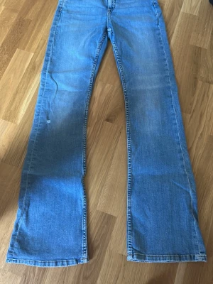 Blå bootcut jeans i klassisk denim - Säljer ett par ljusblå bootcut jeans i klassisk denim med utsvängda ben och normalhög midja. Jeansen har fem fickor och dragkedja med knapp framtill. Perfekta för dig som gillar retrostil och vill ha ett par bekväma jeans till vardags. (jeansen har ett litet hål)