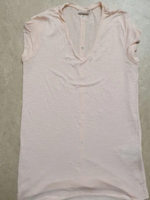 T-shirt - Ljusrosa t-shirt i tunn bomull med v-ringning och korta ärmar. T-shirten har dekorativa knappar längs ryggen och en avslappnad passform. Perfekt för varma dagar och enkel att matcha med jeans eller kjol.