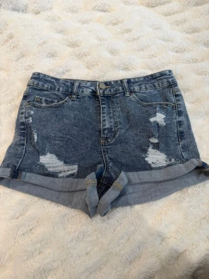 Blåa denimshorts med slitningar - Snygga blåa jeansshorts med slitna detaljer och uppvikta benslut. Klassisk femficksmodell med knapp och dragkedja framtill. Perfekta för sommaren och ger en avslappnad vibe.