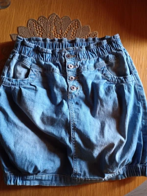 Blå jeanskjol med knappar - Säljer en blå jeanskjol med resår i midjan och flera knappar framtill. Kjolen har två fickor fram och två bak, samt en avslappnad passform. Perfekt till vardags för en avslappnad och cool stil.