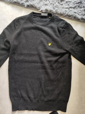 Svart tröja från Lyle & Scott - Klassisk svart tröja från Lyle & Scott med rund halsringning och den ikoniska gula örnloggan broderad på bröstet. Tröjan har ribbade muddar vid ärmslut och nederkant. Perfekt för en stilren och enkel look.