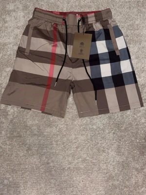 Rutig Burberry shorts med snörning - Snygga shorts från Burberry med klassiskt rutigt mönster i beige, brunt, svart, vitt och rött. Shortsen har elastisk midja med snörning och är perfekta för en trendig och avslappnad look. Ikonisk design som sticker ut.