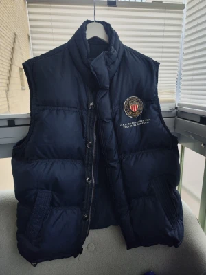 Mörkblå dunväst från Polo Jeans Co. Ralph Lauren Storlek XL - Snygg mörkblå dunväst från Polo Jeans Company med hög krage och broderad patch på bröstet. Västen har knappar och dragkedja framtill samt två fickor med ribbade detaljer. Perfekt för lager på lager och streetwear-stil. Vintage Retro Väst! Storlek XL. Har inte använts. 